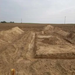 Wykonane wykopy w gruncie pod przyszłe fundamenty budynku, widoczne ślady opon i pryzmy ziemi.