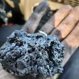 Dłoń w roboczych rękawicach trzyma bryłę sadzy o nieregularnej strukturze, widoczny pierścionek na palcu, w tle elementy instalacji kominowej.