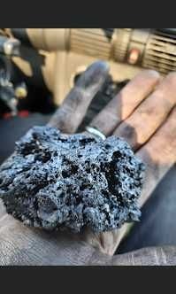 Dłoń w roboczych rękawicach trzyma bryłę sadzy o nieregularnej strukturze, widoczny pierścionek na palcu, w tle elementy instalacji kominowej.