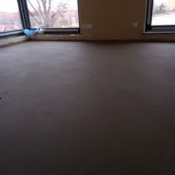 Świeżo wylana, szara posadzka w pomieszczeniu z dużymi oknami, widoczne gniazdka elektryczne na ścianie.