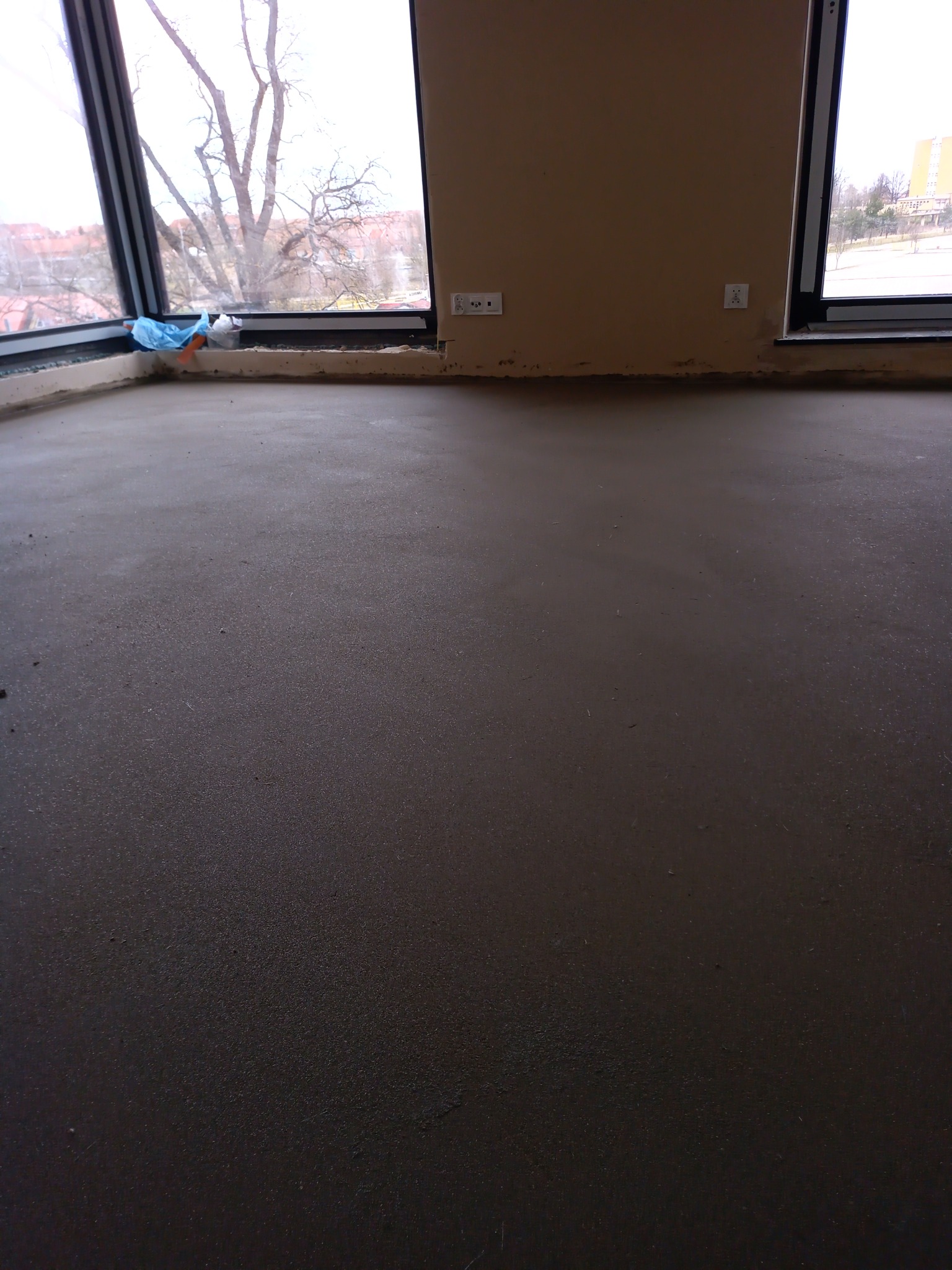 Świeżo wylana, szara posadzka w pomieszczeniu z dużymi oknami, widoczne gniazdka elektryczne na ścianie.