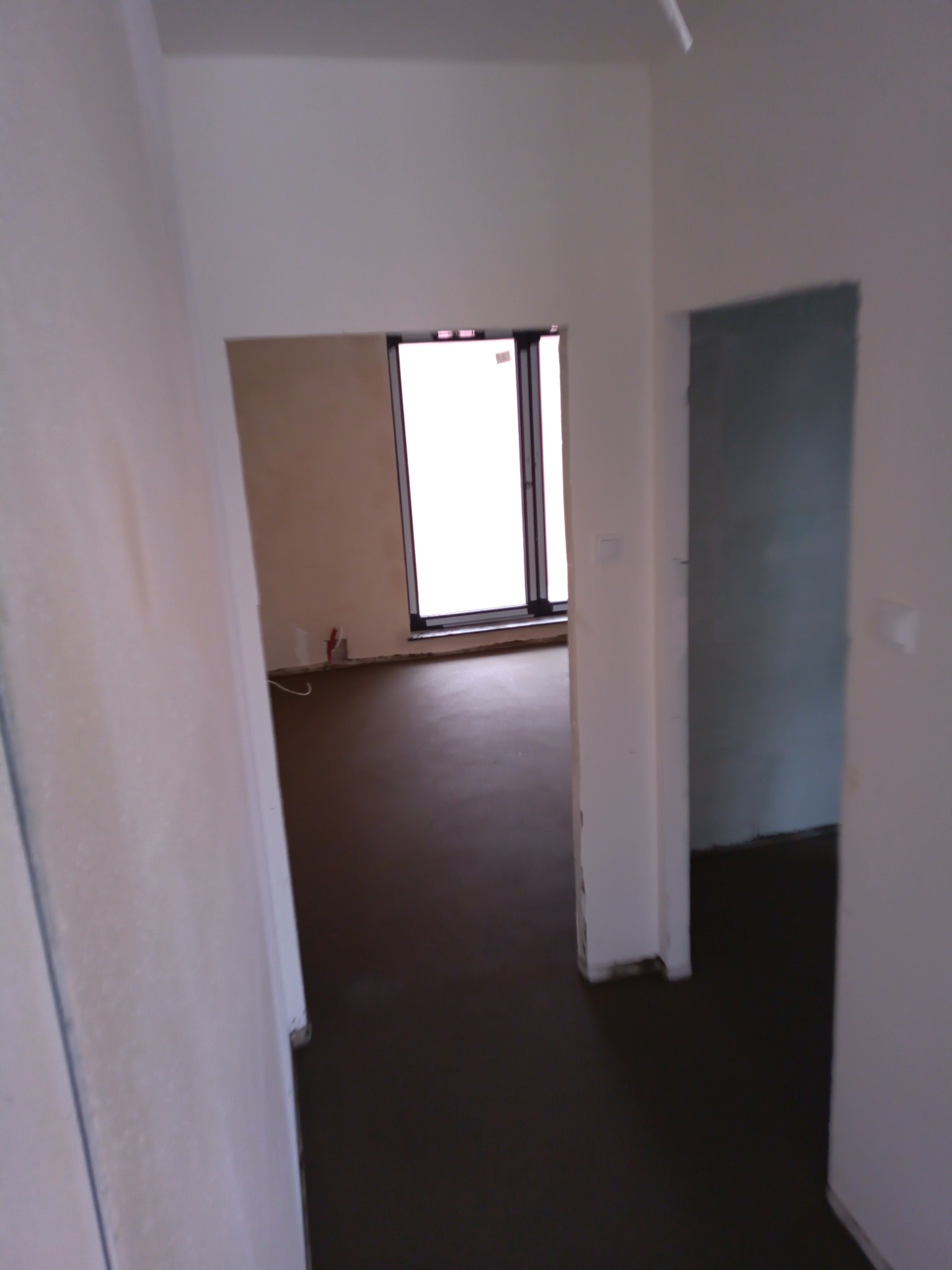 Świeżo wylana, ciemna posadzka jastrychowa w surowym stanie, widok z korytarza na pokój z oknem, widoczne ślady po pracach budowlanych na ścianach.