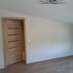 Jasne panele podłogowe ułożone w pokoju z drewnianą ościeżnicą i nowoczesną lampą sufitową, widoczny fragment nieułożonych paneli.