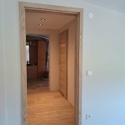Zbigniew Banasiak - Widok na korytarz z drewnianą podłogą i ościeżnicą w kolorze jasnego drewna, widoczne drzwi i fragment szafy w tle, białe ściany z włącznikiem i gniazdkiem elektrycznym.