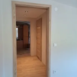 Widok na korytarz z drewnianą podłogą i ościeżnicą w kolorze jasnego drewna, widoczne drzwi i fragment szafy w tle, białe ściany z włącznikiem i gniazdkiem elektrycznym.