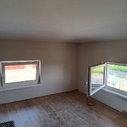 Zbigniew Banasiak - Pokój po remoncie, z nową podłogą z paneli drewnopodobnych, dwa okna z białymi ramami, ściany pomalowane na beżowo, widoczne listwy przypodłogowe i narzędzia.