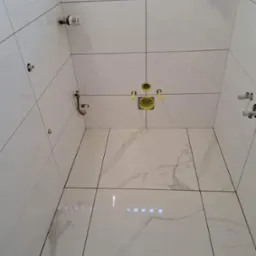 Wnętrze kabiny prysznicowej wyłożonej białymi płytkami imitującymi marmur, z widocznymi instalacjami hydraulicznymi i grzewczymi przed montażem armatury.