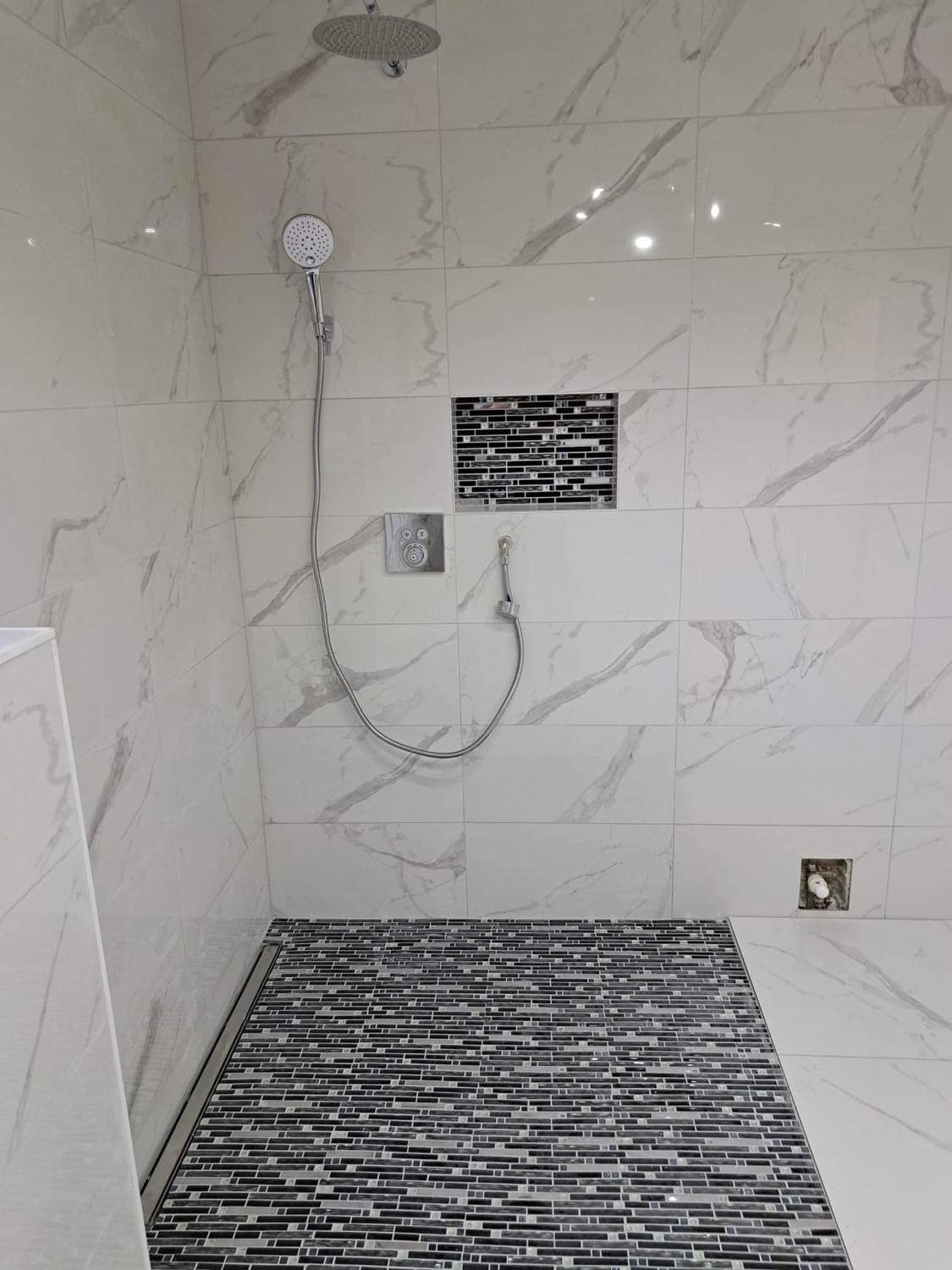 Nowoczesny prysznic z marmurowymi płytkami ściennymi, mozaikową podłogą i wnęką, deszczownicą sufitową i słuchawką prysznicową.