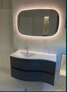 Nowoczesna łazienka z umywalką nablatową na szafce z szufladami o falistym froncie w kolorze ciemnego drewna, podświetlane lustro o nieregularnym kształcie, białe płytki imitujące marmur...