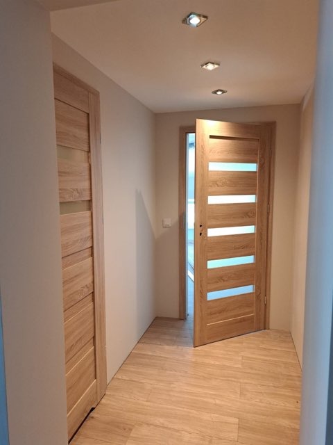 Jasne panele podłogowe w korytarzu z dwiema drewnianymi drzwiami z przeszkleniami, oświetlenie punktowe w suficie.