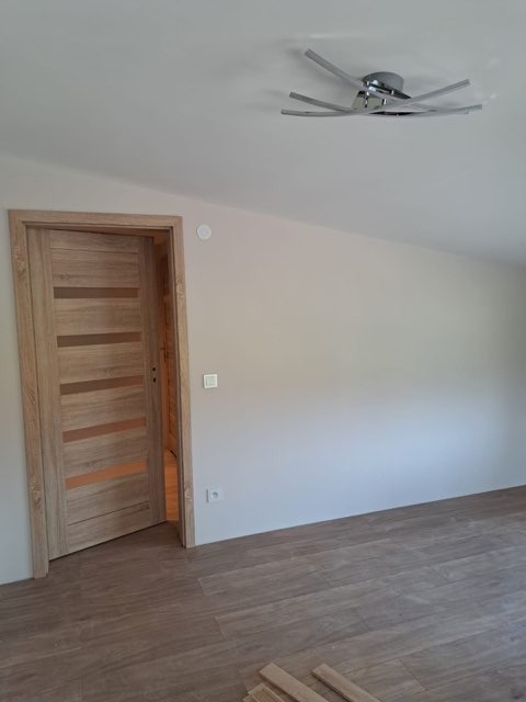 Jasne panele podłogowe ułożone w pokoju z drewnianą ościeżnicą i nowoczesną lampą sufitową, widoczny fragment nieułożonych paneli.