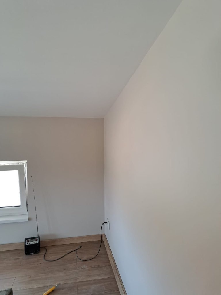 Świeżo pomalowane, jasne pomieszczenie z drewnopodobną podłogą, widoczne gniazdko elektryczne, radio i fragment okna.