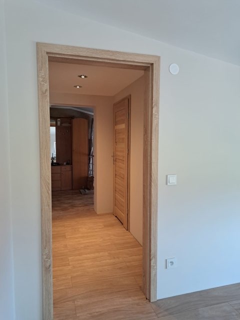 Widok na korytarz z drewnianą podłogą i ościeżnicą w kolorze jasnego drewna, widoczne drzwi i fragment szafy w tle, białe ściany z włącznikiem i gniazdkiem elektrycznym.