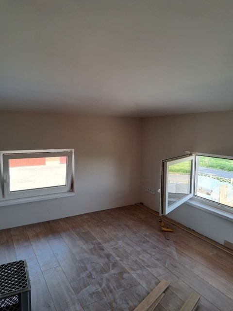 Pokój po remoncie, z nową podłogą z paneli drewnopodobnych, dwa okna z białymi ramami, ściany pomalowane na beżowo, widoczne listwy przypodłogowe i narzędzia.