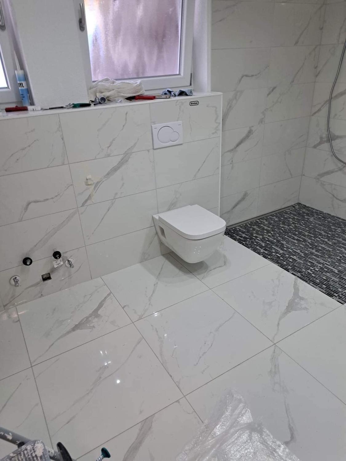 Nowoczesna łazienka z białymi płytkami imitującymi marmur, podwieszana toaleta i mozaiką w strefie prysznica, widok z góry na świeżo wykończone wnętrze.