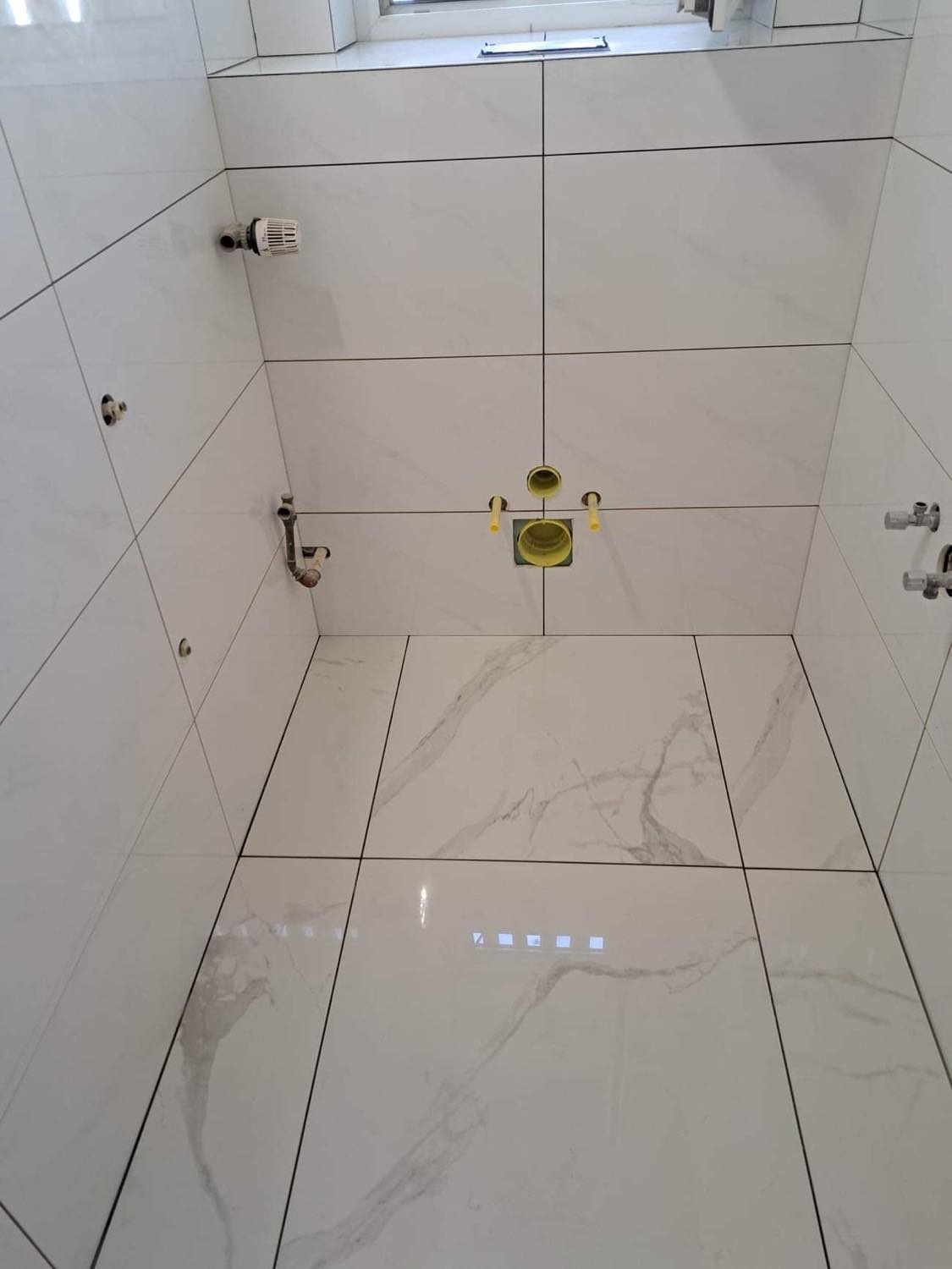 Wnętrze kabiny prysznicowej wyłożonej białymi płytkami imitującymi marmur, z widocznymi instalacjami hydraulicznymi i grzewczymi przed montażem armatury.