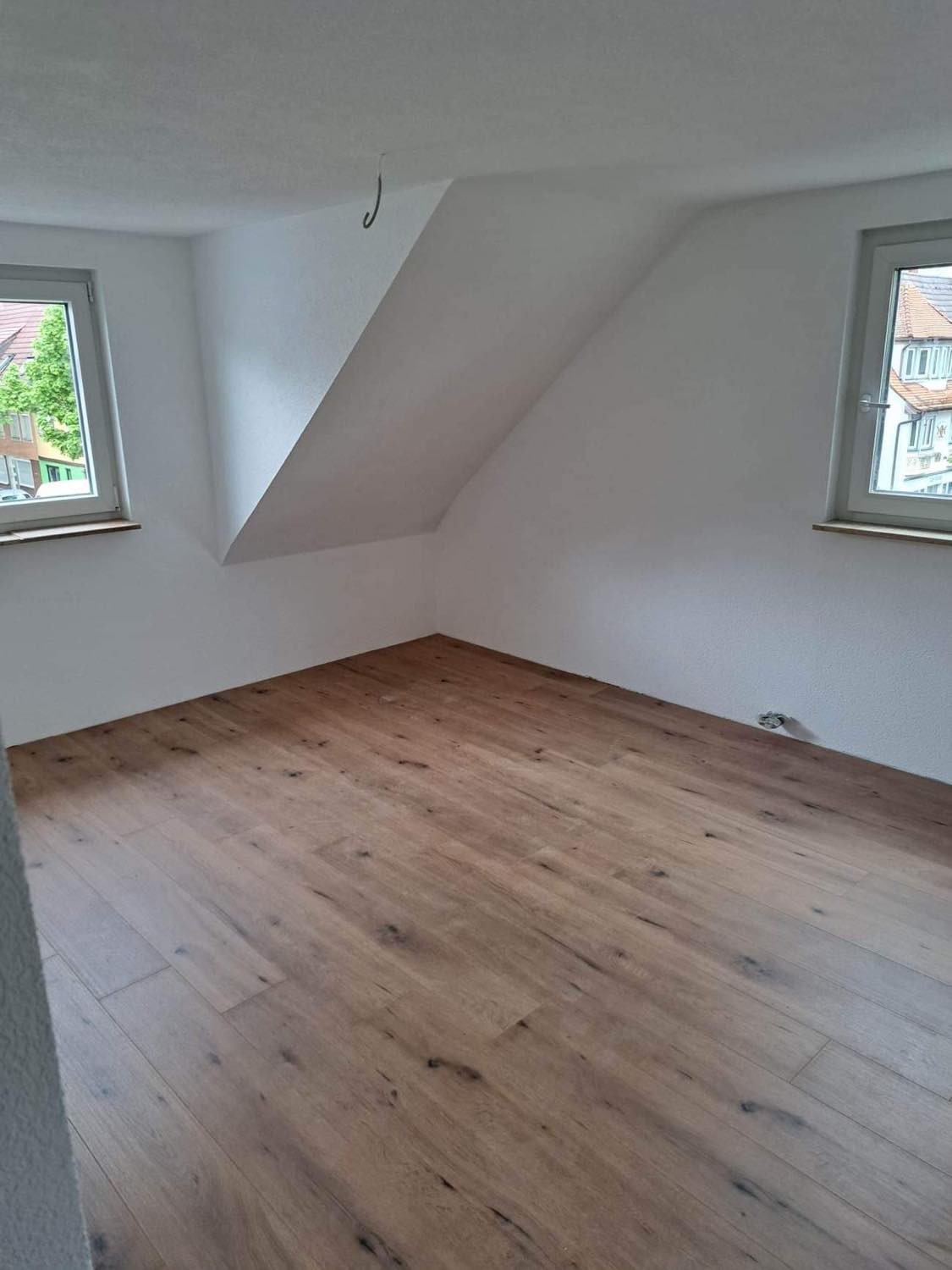 Poddasze po remoncie z nową, jasną podłogą z paneli drewnopodobnych, białymi ścianami i dwoma oknami z widokiem na zieleń.