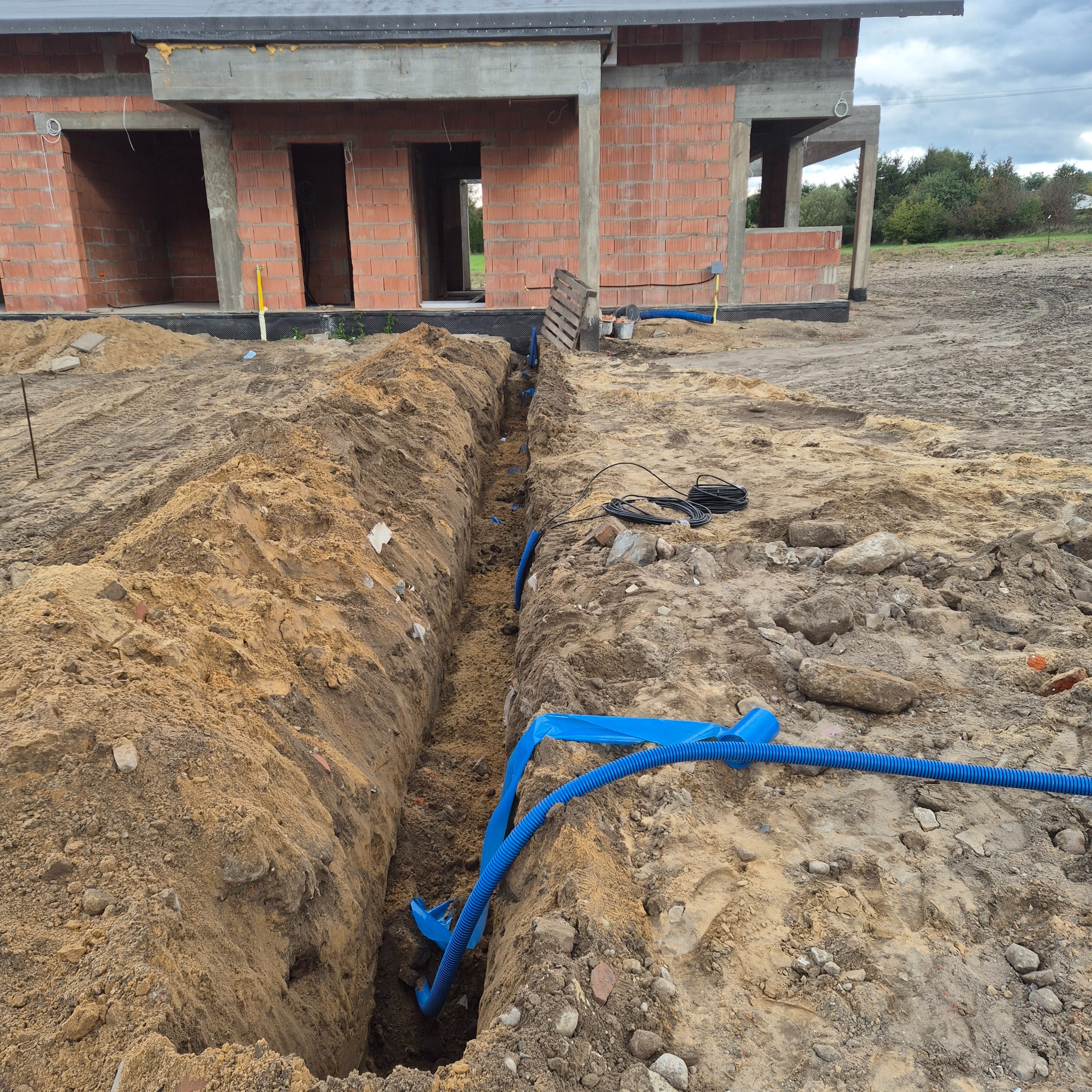 Wykop z ułożonymi niebieskimi rurami kablowymi w ziemi, widoczny budynek w stanie surowym otwartym z cegły i z wystającymi przewodami elektrycznymi.