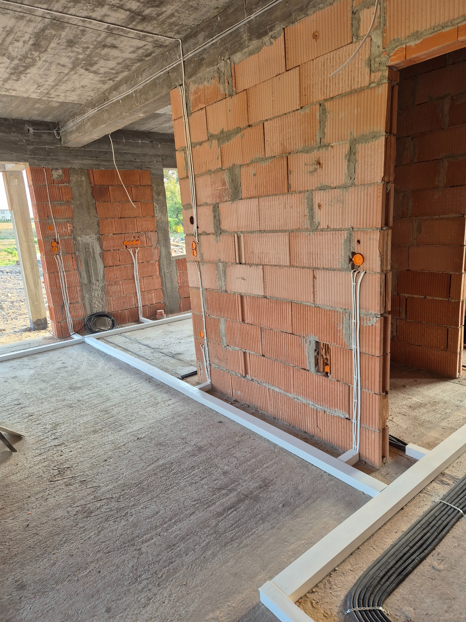 Wewnętrzne ściany z cegły dziurawki w trakcie budowy, widoczne przewody elektryczne prowadzone wzdłuż ścian i pod sufitem, pomarańczowe puszki instalacyjne, białe listwy przypodłogowe i wiązka rur...