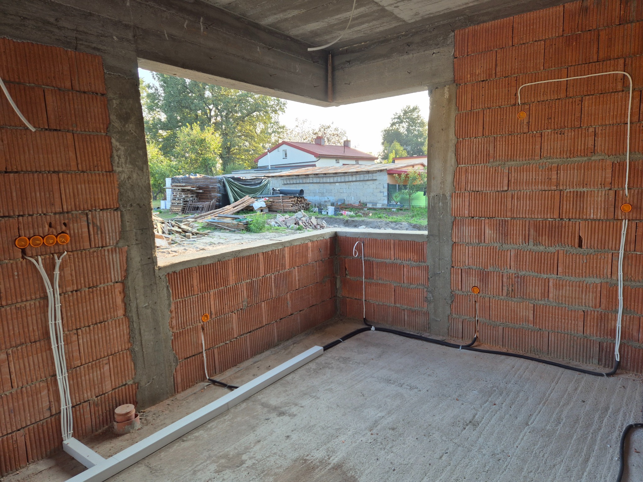 Wewnętrzne ściany z czerwonej cegły z widocznymi przewodami elektrycznymi w rurkach ochronnych, wychodzącymi z pomarańczowych puszek instalacyjnych, z widokiem na zewnątrz przez niezabudowany otwór...