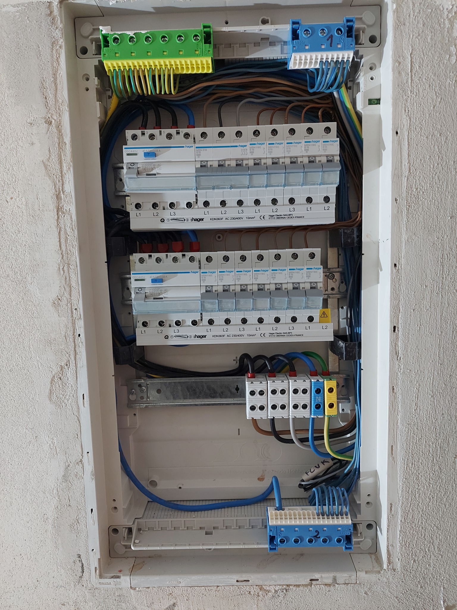 Rozdzielnica elektryczna Hager w trakcie montażu, widoczne zabezpieczenia nadprądowe, różnicowoprądowe, listwy zaciskowe w kolorach zielonym, niebieskim i żółto-niebieskim, przewody w różnych...