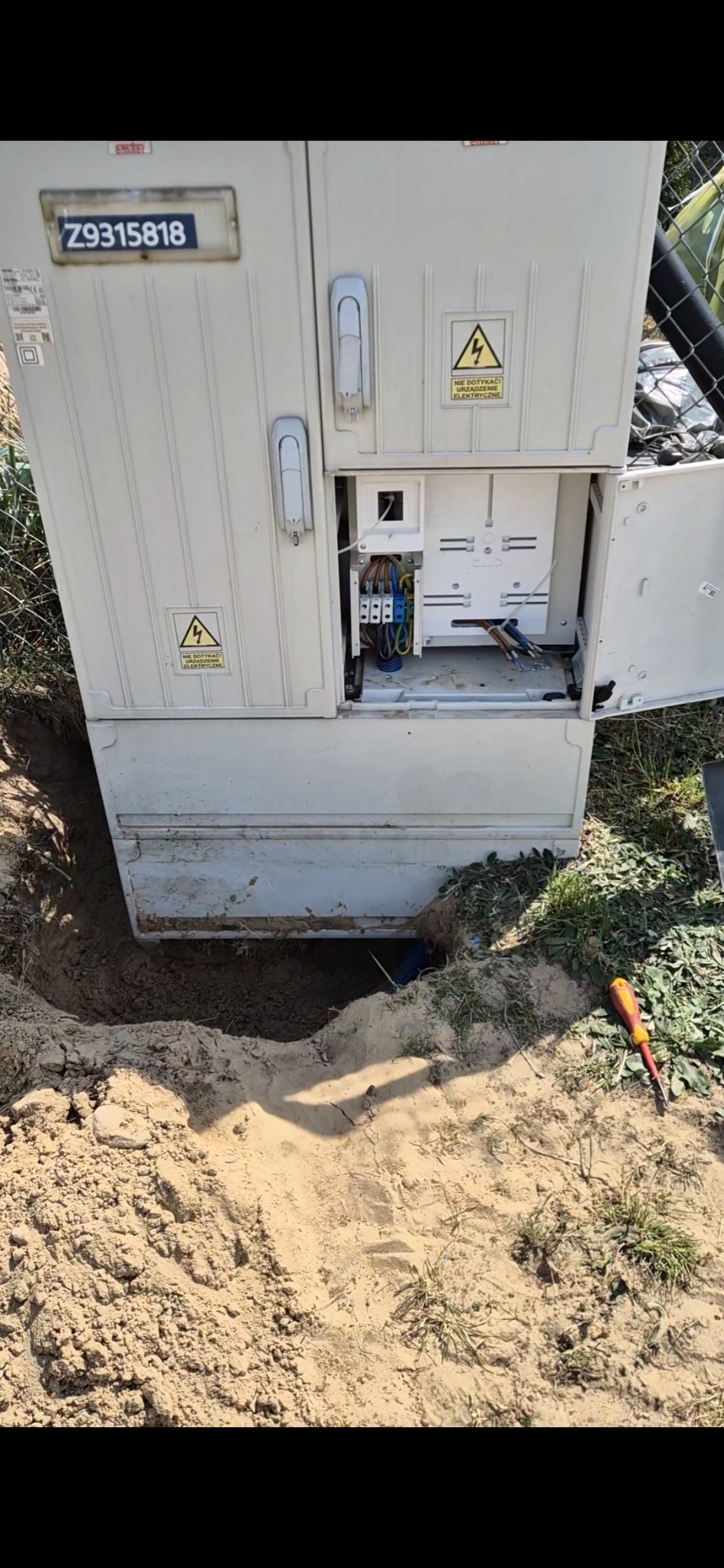 Otwarta skrzynka elektryczna z numerem seryjnym Z9315818, częściowo zakopana w ziemi, obok leży śrubokręt. Widoczne kable wewnątrz.