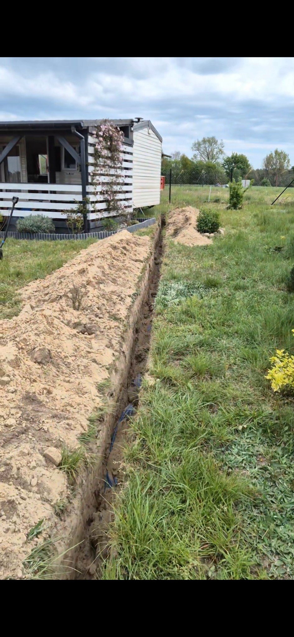 Wykop w ziemi z ułożonym kablem w pobliżu domu z białym ogrodzeniem. Widoczne świeżo usypane brzegi wykopu i trawa. Prace ziemne przed zakopaniem kabla.