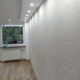 Ściana w pokoju po szpachlowaniu i malowaniu, widoczna tekstura, oświetlenie punktowe w suficie, podłoga z paneli, okno z widokiem na zieleń na zewnątrz.