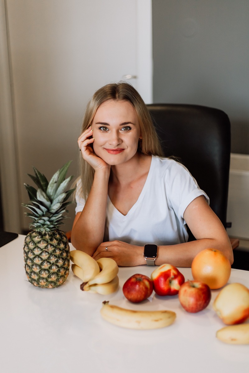Uśmiechnięta blondynka siedzi za białym stołem z ananasem, bananami, jabłkami i grejpfrutem, opierając brodę na dłoni z zegarkiem.