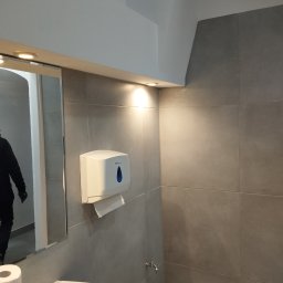 Mikołaj Mariusz Chudy - Szara łazienka z umywalką, dozownikiem ręczników papierowych i lustrem, w którym odbija się osoba w ciemnym ubraniu.