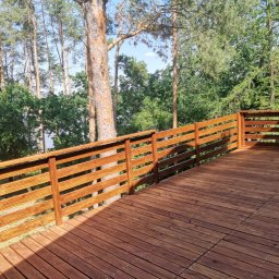 Mikołaj Mariusz Chudy - Nowo wybudowany drewniany taras z brązową balustradą, otoczony zielenią drzew i krzewów.