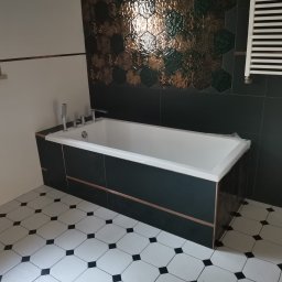 Mikołaj Mariusz Chudy - Nowoczesna łazienka z wanną obudowaną ciemnymi płytkami z miedzianą listwą, heksagonalne płytki dekoracyjne na ścianie i biało-czarna podłoga w romby.