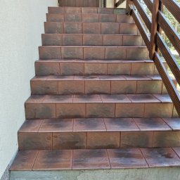 Mikołaj Mariusz Chudy - Zewnętrzne schody wejściowe wyłożone brązowymi płytkami imitującymi kamień, z drewnianą balustradą po prawej stronie, prowadzące do ciemnobrązowych drzwi wejściowych.