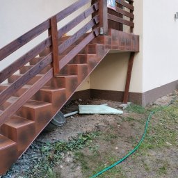 Mikołaj Mariusz Chudy - Schody zewnętrzne wyłożone brązowymi płytkami z drewnianą balustradą, widoczny numer domu 3A, teren wokół schodów częściowo zagospodarowany, leży wiadro i fragment styropianu, wąż ogrodowy na ziemi.