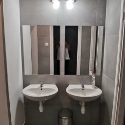 Mikołaj Mariusz Chudy - Szara łazienka z dwoma umywalkami, lustrami z oświetleniem i srebrnym koszem na śmieci. W lustrach odbija się osoba robiąca zdjęcie.