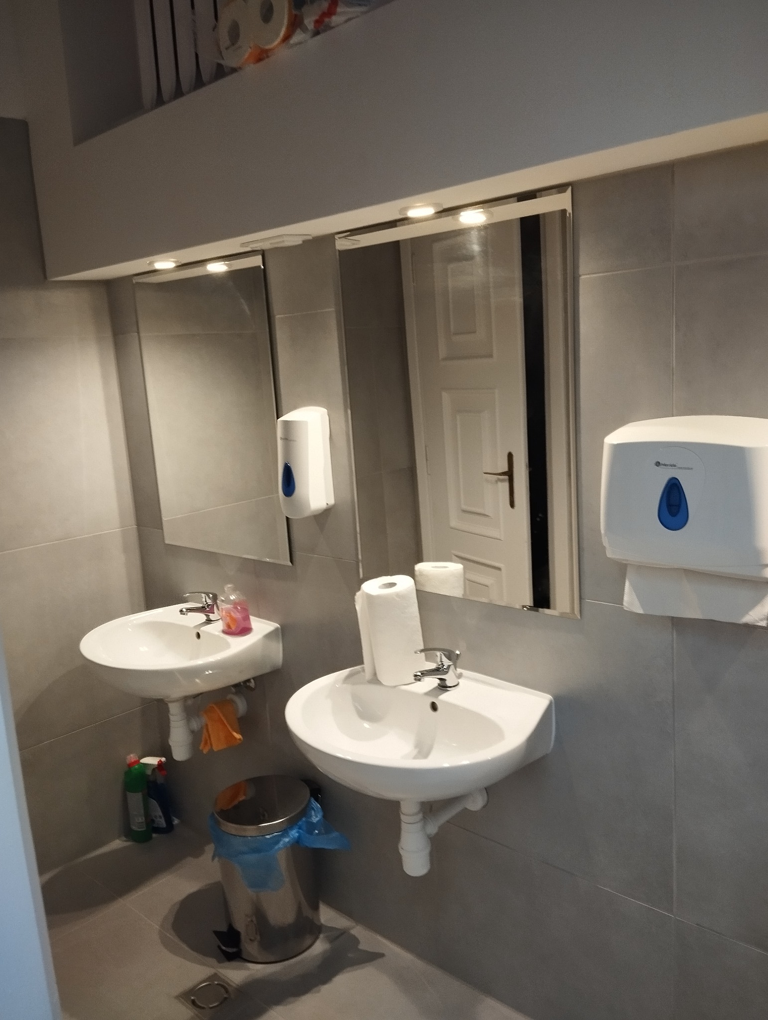 Umywalki ceramiczne zamontowane na szarej ścianie, z dozownikiem mydła i papieru, widoczny kosz na śmieci z niebieską folią, oraz odbicie białych drzwi w lustrze.