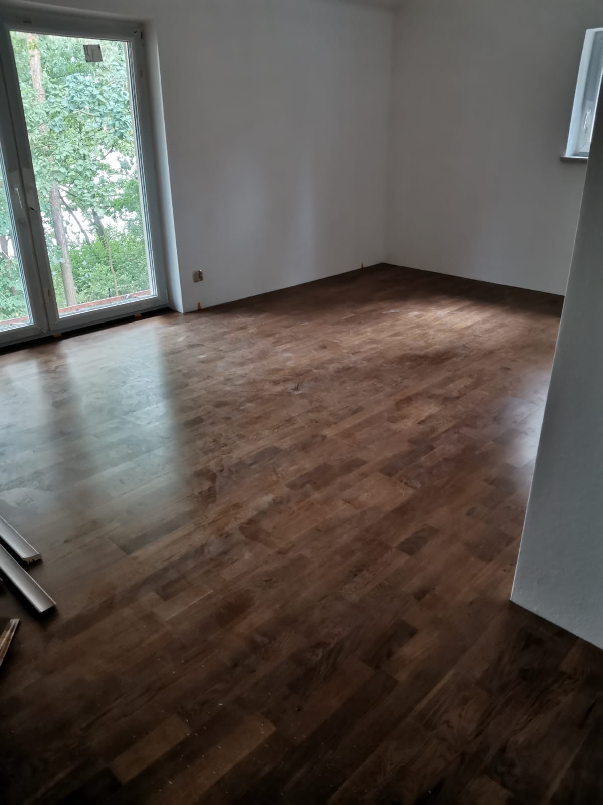 Pokój po remoncie z nową, błyszczącą, drewnianą podłogą w odcieniach brązu, widok na zieleń za oknem, listwy przypodłogowe częściowo ułożone przy ścianie.