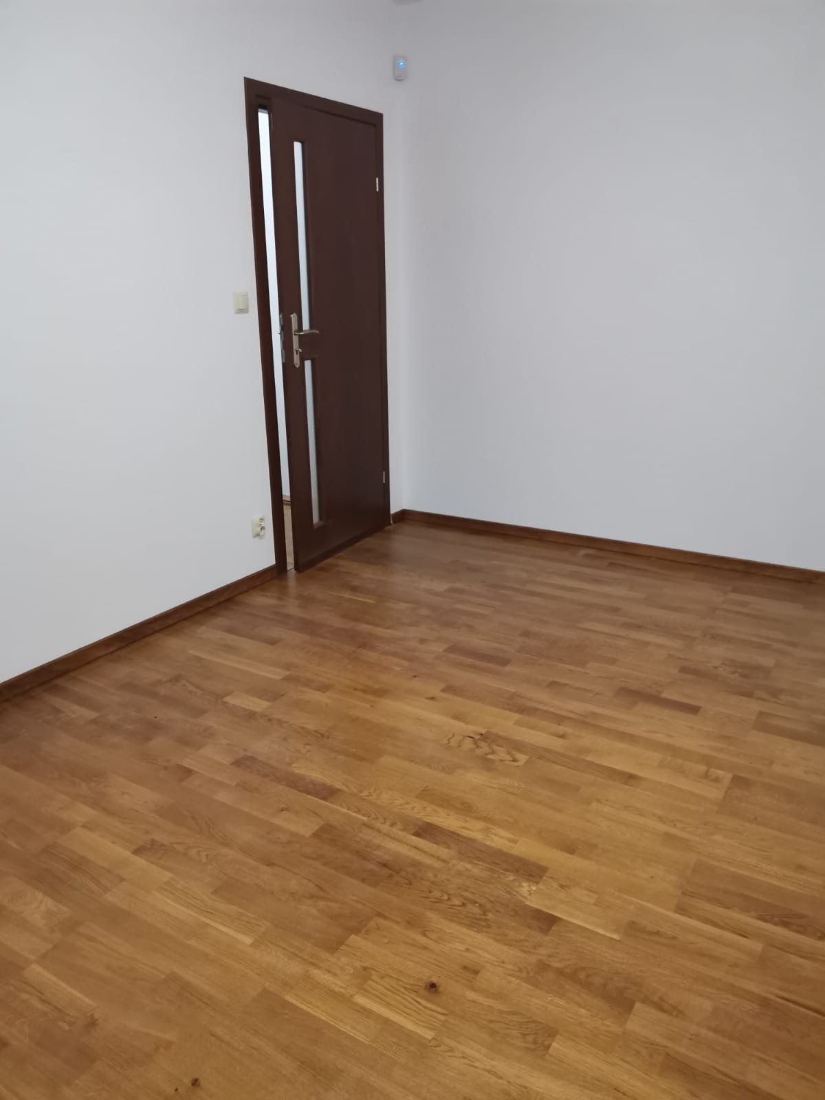 Pusty pokój z drewnianą podłogą i brązowymi drzwiami z przeszkleniem, świeżo po położeniu parkietu.