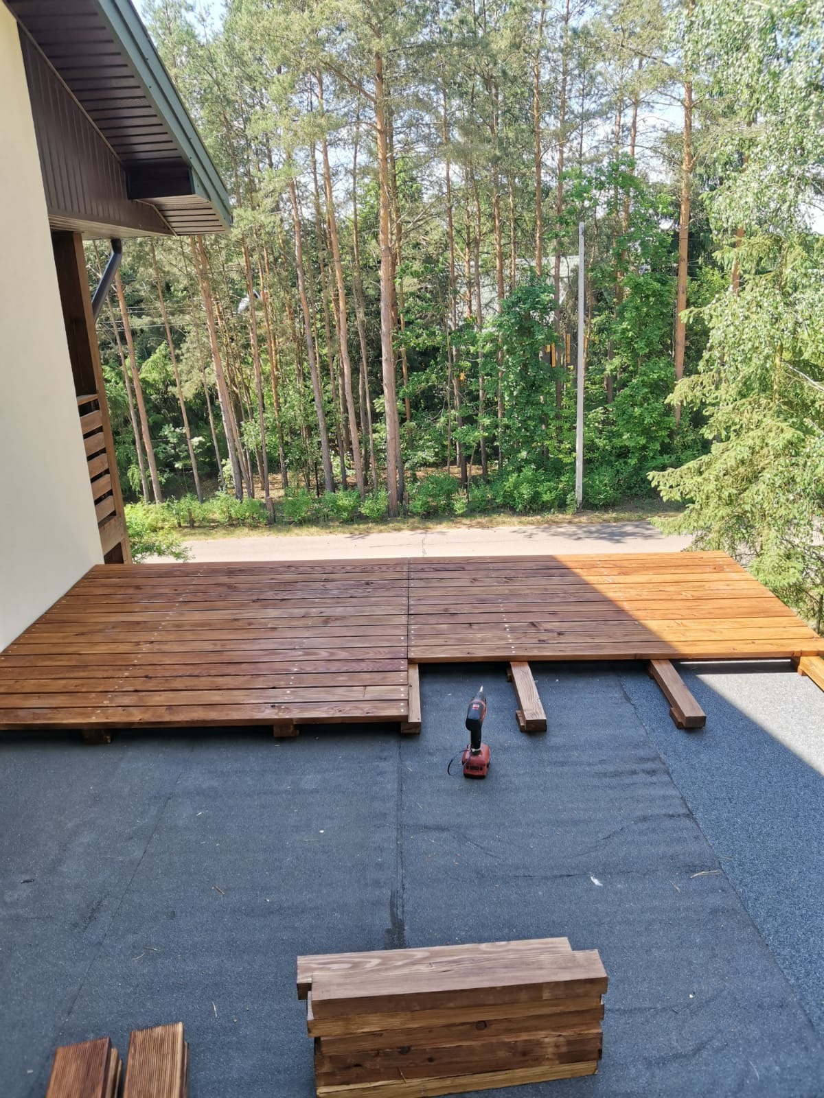 Montaż drewnianego tarasu przy domu z użyciem wkrętarki, widoczne deski w kolorze brązowym, legary i podłoże z papy, w tle las.