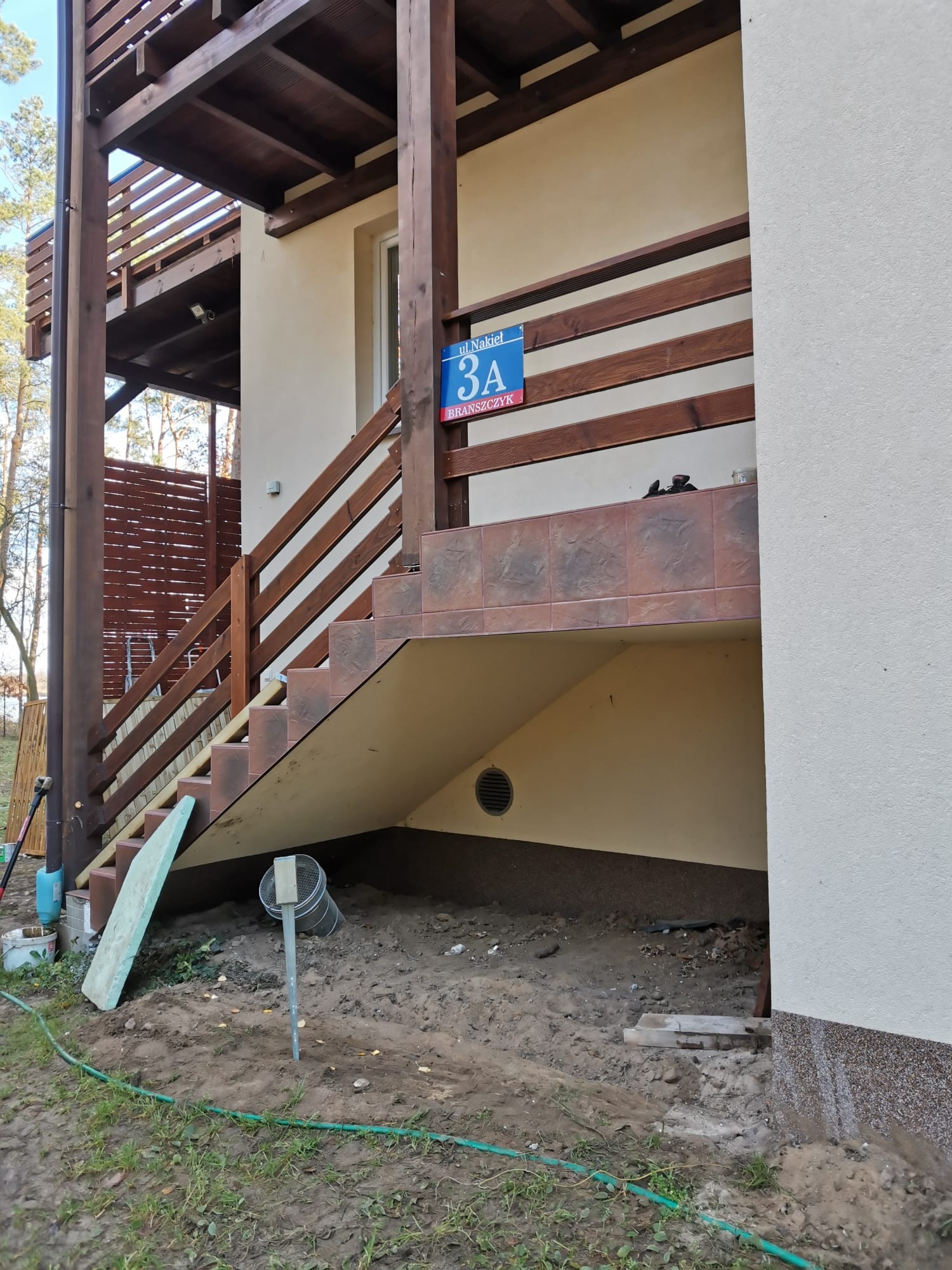 Zewnętrzne schody z brązowymi płytkami prowadzące na drewniany balkon, widoczny adres na niebieskiej tabliczce, teren wokół schodów w trakcie prac ziemnych.