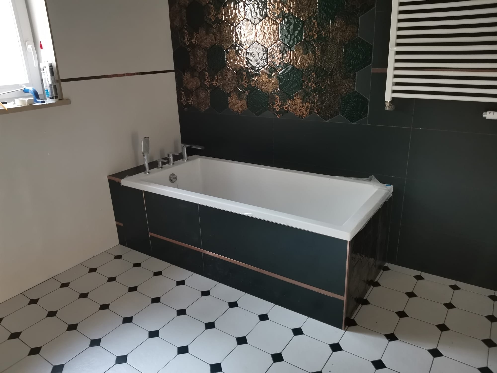 Nowoczesna łazienka z wanną obudowaną ciemnymi płytkami z miedzianą listwą, heksagonalne płytki dekoracyjne na ścianie i biało-czarna podłoga w romby.