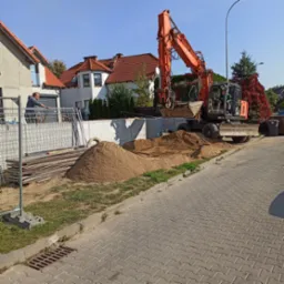 Pomarańczowa koparka Hitachi na placu budowy, obok sterty piasku i fundamentów, z pracownikiem opierającym się o ogrodzenie w tle.