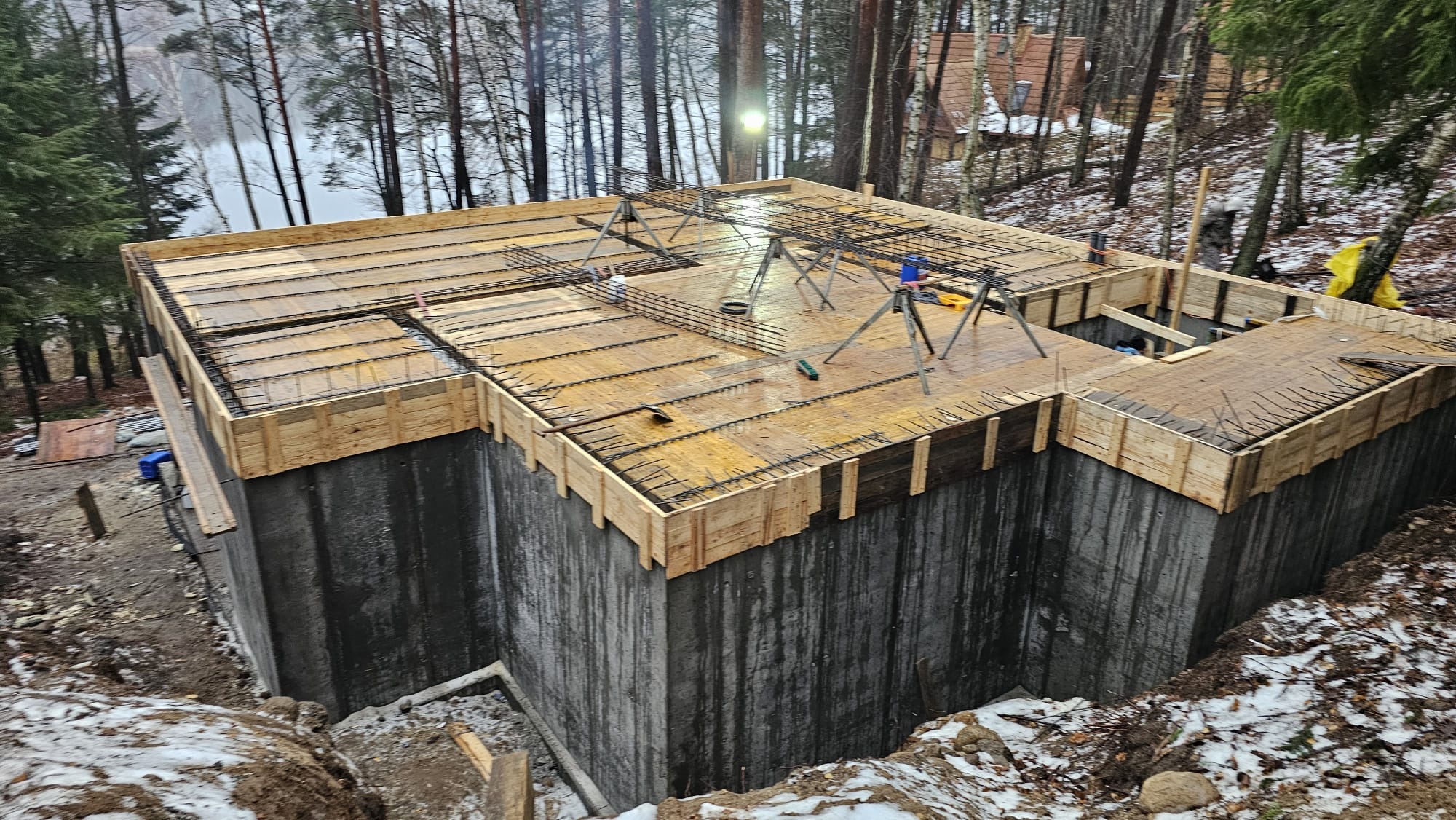 Betonowy fundament z drewnianym szalunkiem i zbrojeniem stalowym, przygotowany do wylania płyty, widok z góry na tle zalesionego terenu w zimowej scenerii.