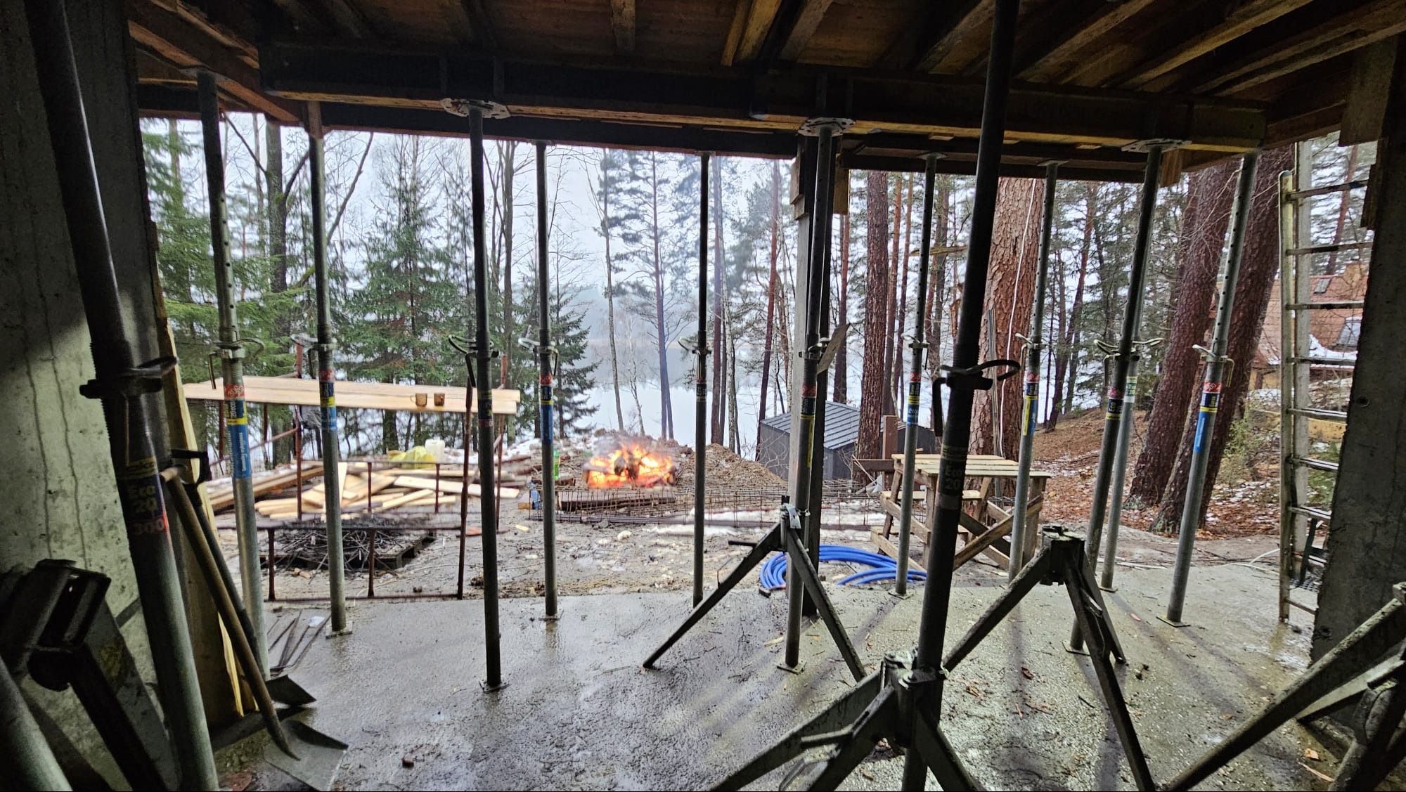 Wnętrze surowego budynku w Olsztynie w trakcie budowy, widoczne podpory stropowe, zbrojenie fundamentów i ognisko palące się na zewnątrz, za drzewami