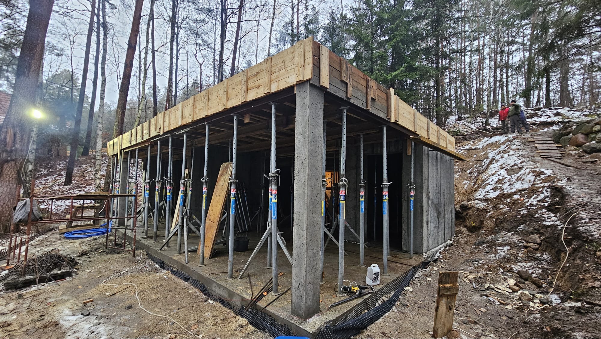 Surowy betonowy szkielet budynku z drewnianym szalunkiem i metalowymi podporami, widok z zewnątrz na tle zalesionego terenu i dwóch osób na wzgórzu.