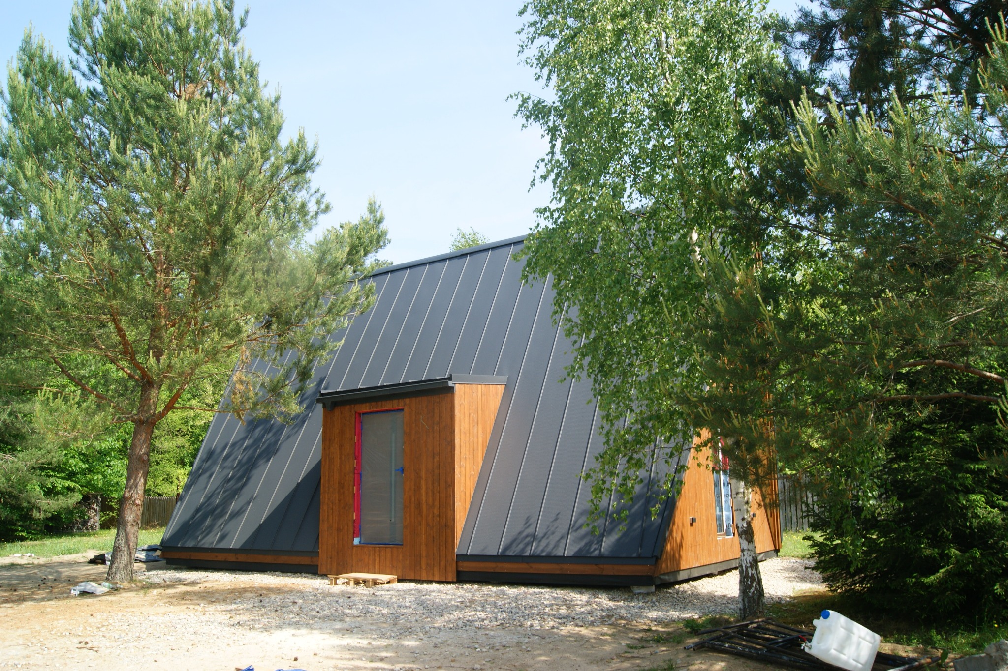Nowoczesny, trójkątny dom z szarą, metalową dachówką, drewnianą elewacją i oknem zabezpieczonym folią, otoczony młodymi drzewami iglastymi i liściastymi na żwirowym podłożu.