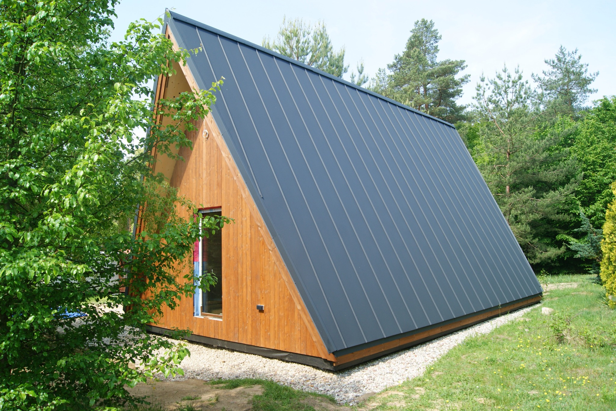 Mały, nowoczesny dom o trójkątnym kształcie z drewnianą elewacją i grafitowym, metalowym dachem, otoczony zielenią.