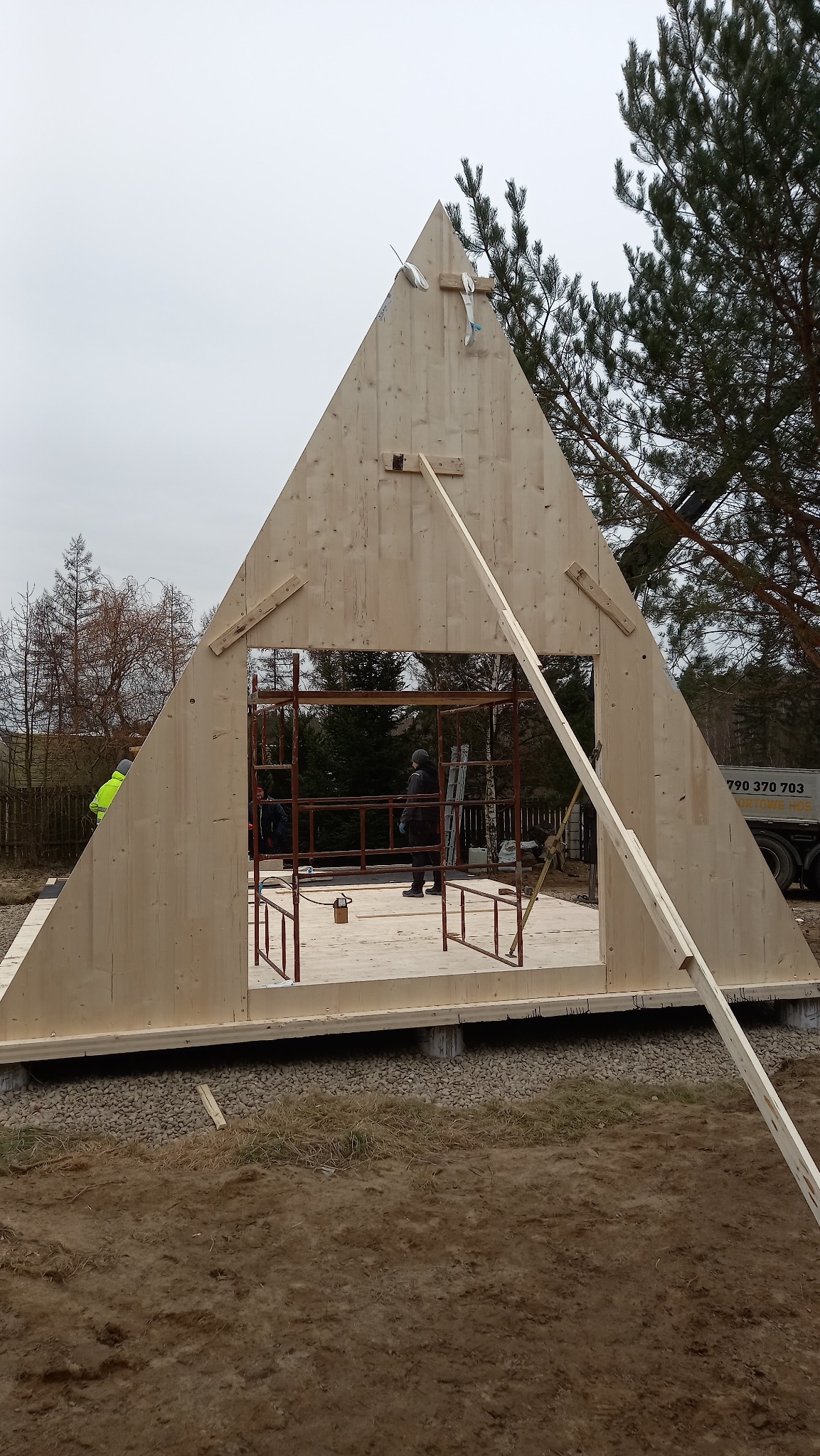 Montaż ściany szczytowej domu szkieletowego na fundamencie, widoczne rusztowania i częściowo zalesione otoczenie.