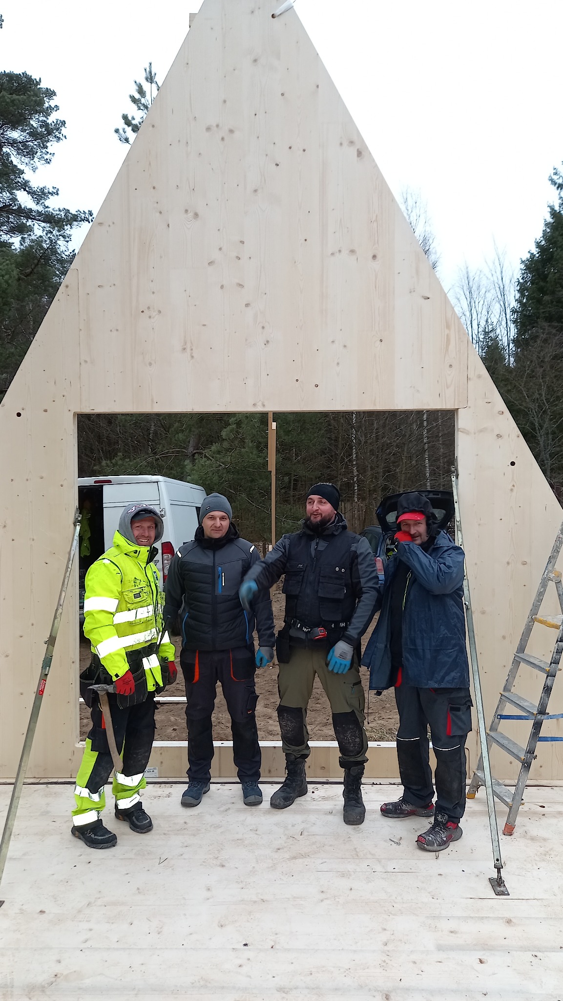 Czterech budowlańców w odzieży roboczej pozuje na tle wznoszonej drewnianej konstrukcji domu o trójkątnym szczycie, z widocznym oknem i drabiną, na placu budowy z lasem w tle.