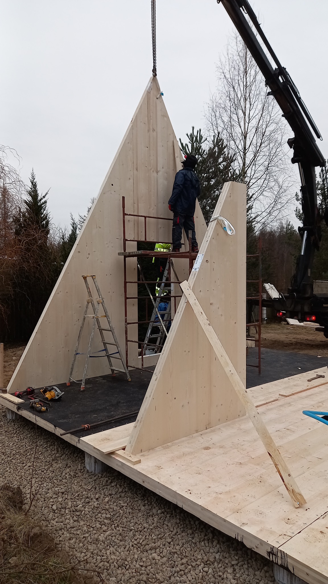 Montaż ściany szkieletowej domu z użyciem dźwigu, pracownik na rusztowaniu, widoczne narzędzia i podłoże z kamieni.